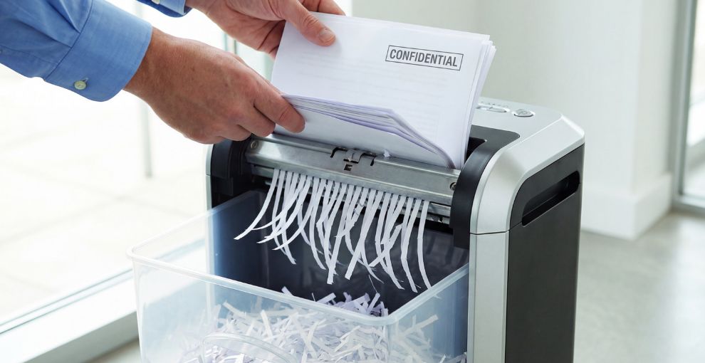 Hands feeding a stack of papers labeled ‘Confidential’ into a paper shredder, with shredded strips collecting below.