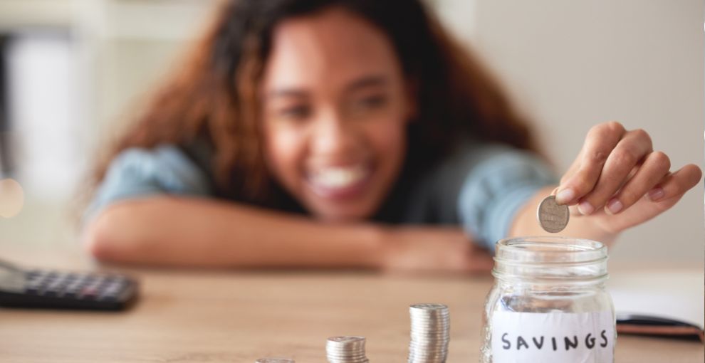 Person placing a coin into a labeled savings jar beside stacks of coins on a table.