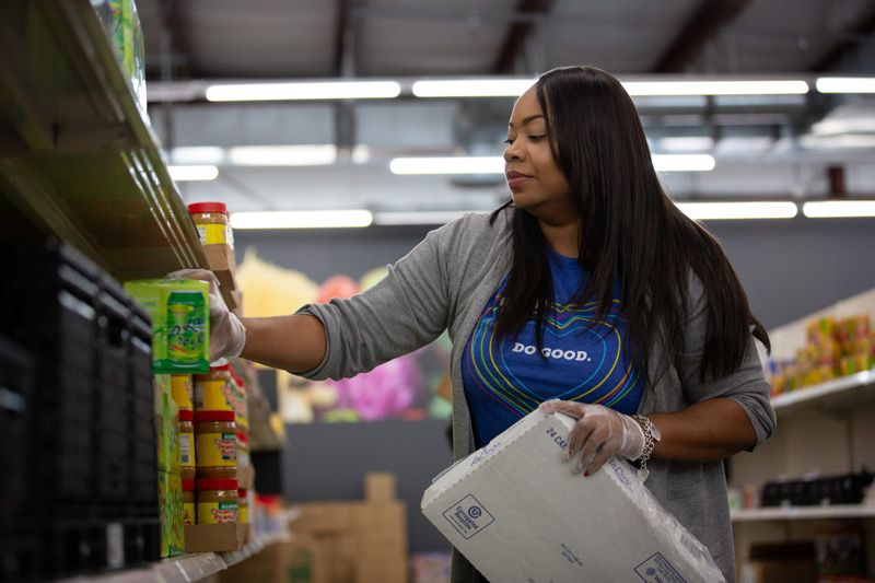 Volunteer stocking jars and canned goods on a food bank shelf while holding a cardboard box.