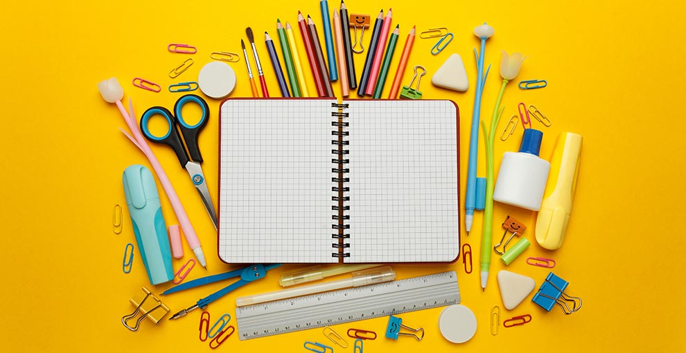 Open notebook surrounded by colorful school supplies including pencils, scissors, ruler, and paper clips on a yellow background.