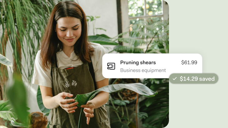 A woman in a white t-shirt and olive green apron with botanical embroidery tends to plants in a bright, plant-filled indoor space. She's holding a large green leaf while surrounded by various houseplants near a window. An overlay shows pruning shears listed as business equipment for $61.99 with $14.29 saved.