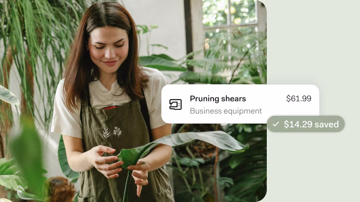 A woman in a white t-shirt and olive green apron with botanical embroidery tends to plants in a bright, plant-filled indoor space. She's holding a large green leaf while surrounded by various houseplants near a window. An overlay shows pruning shears listed as business equipment for $61.99 with $14.29 saved.