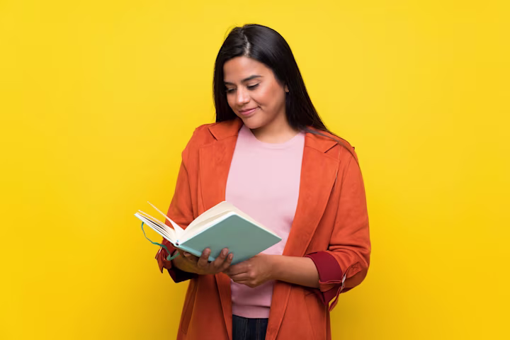 A teenage girl stands in front of a yellow background and reads a book
