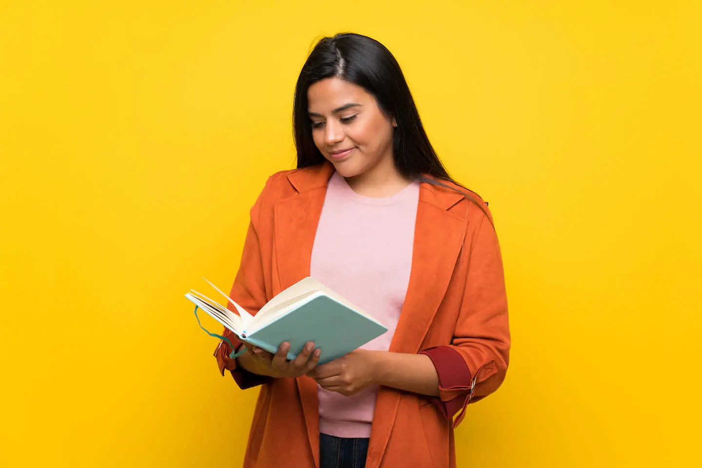 A teenage girl stands in front of a yellow background and reads a book