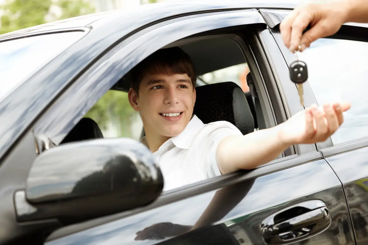 A young teen sits in the driver's seat of a new car and reaches out the window for the key