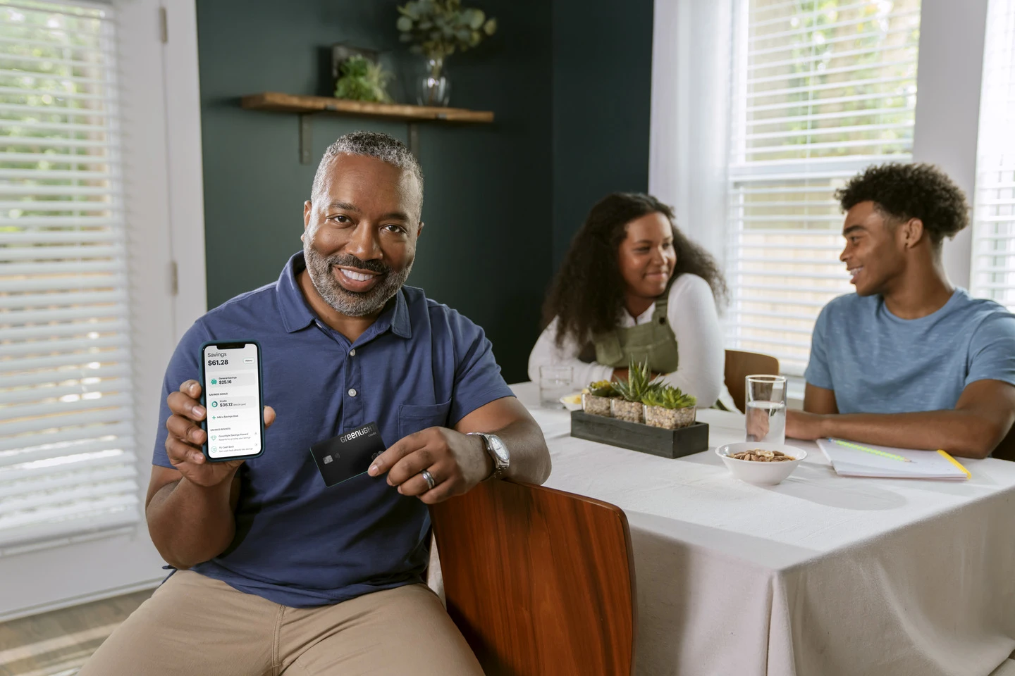 Father showing Greenlight mobile app and debit card with teens in the background