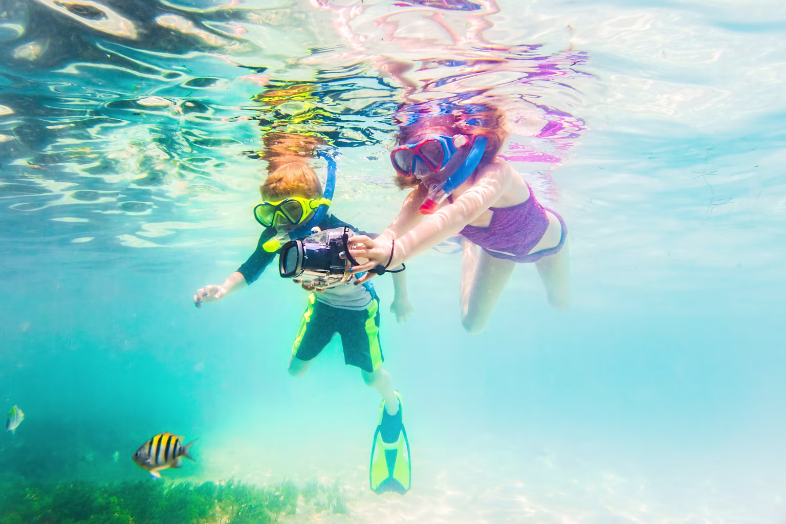 A boy and girl with snorkeling gear on and swimming in the water with a fish.