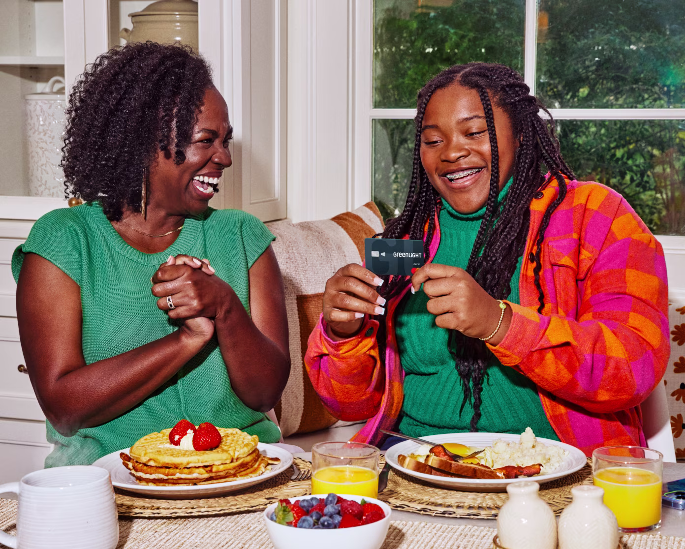 Two women sharing a joyful breakfast moment, one holding a Greenlight card, with pancakes and fruit on the table.
