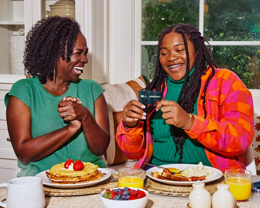 Two women sharing a joyful breakfast moment, one holding a Greenlight card, with pancakes and fruit on the table.