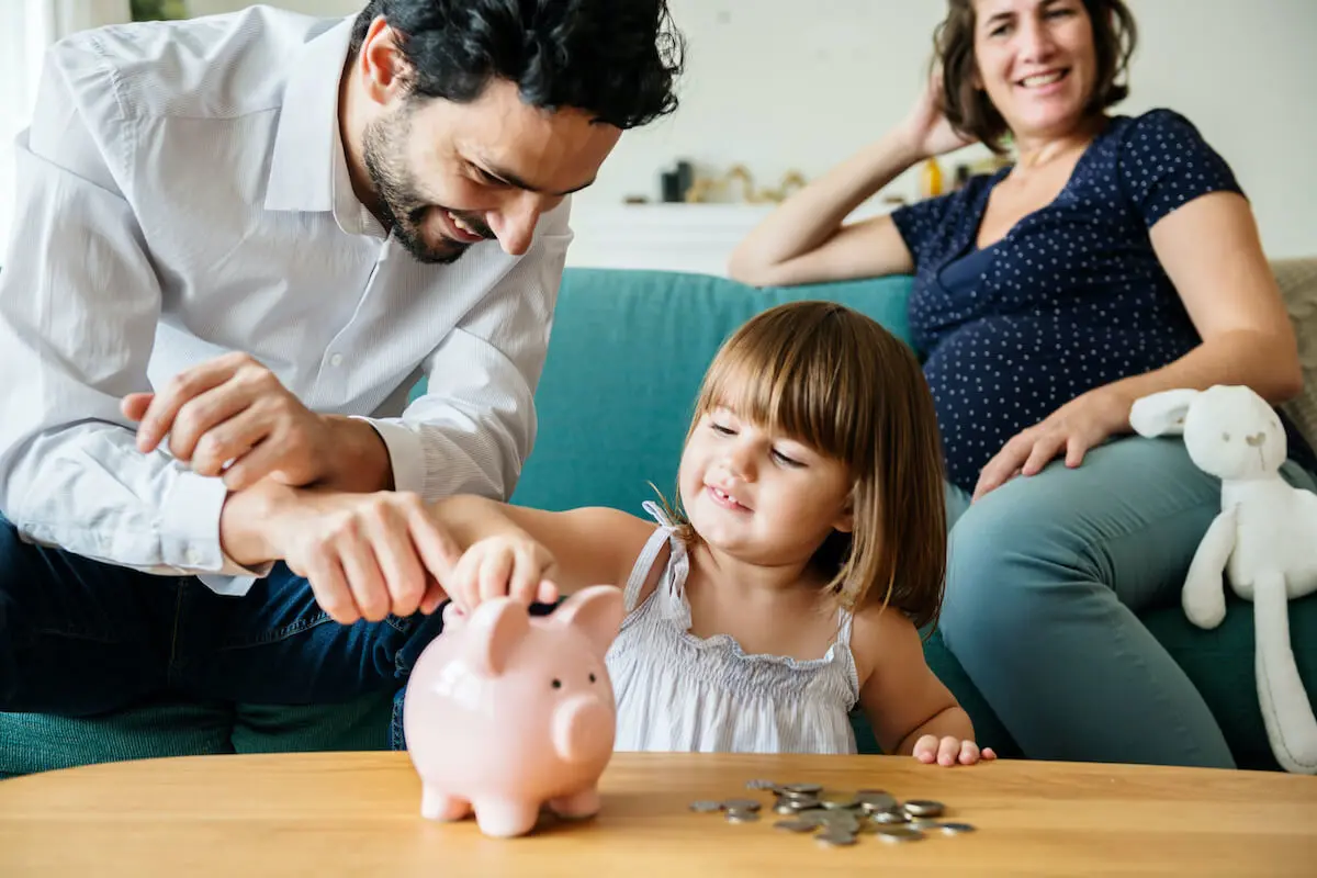 A dad helps a little girl put coins in a piggy bank while the mom smiles in the background