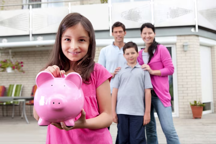 A young girls smiles and holds a piggy bank while her family stands in the background