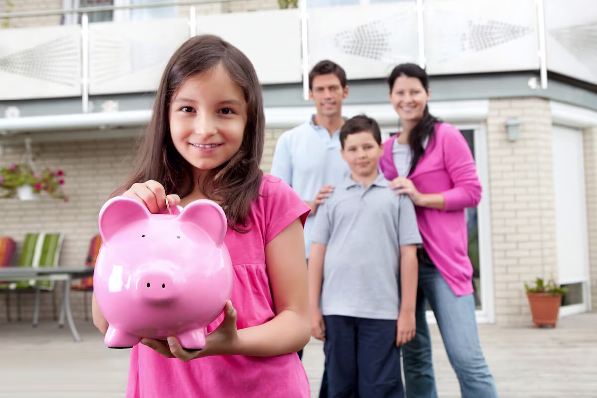 A young girls smiles and holds a piggy bank while her family stands in the background