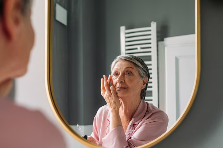 Older woman putting make up on while looking in a mirror.