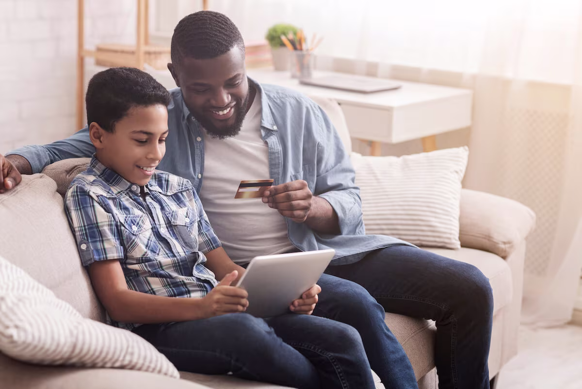 A father is holding a credit card for teens while his son looks at a tablet.