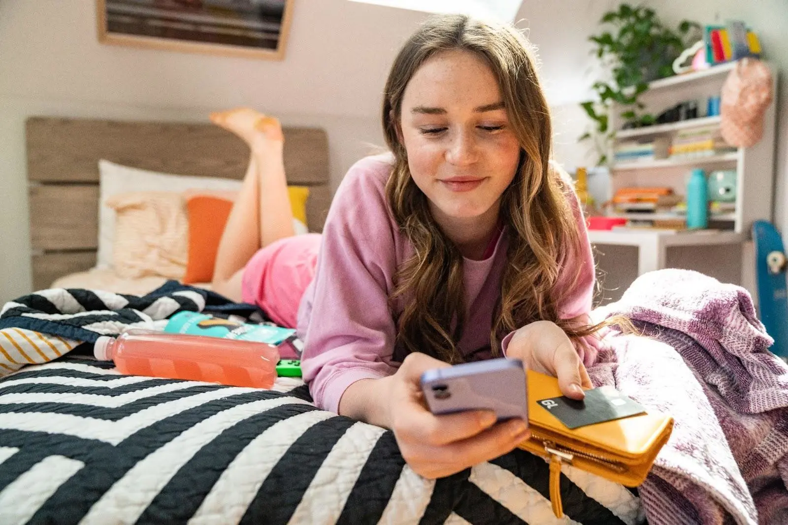 A girl in her room on her phone holding a credit card and wallet.