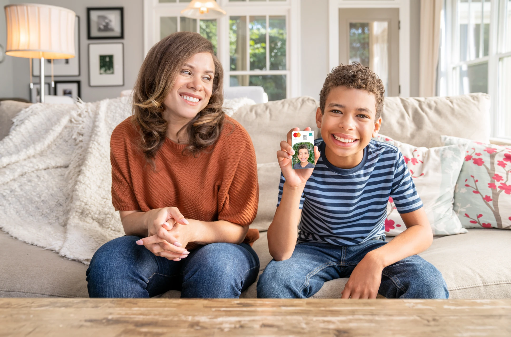 Young boy sitting on couch holding custom Greenlight debit card while mom sits next to him and watches proudly