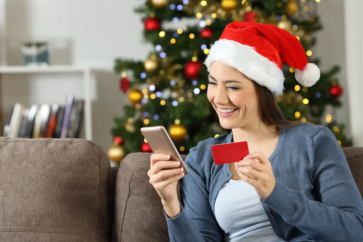 A woman in a Santa hat sits on her couch and checks the balance in her Christmas savings account on her cellphone