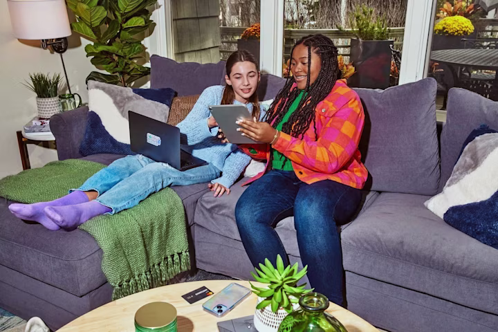 Two girls on a couch with a tablet and a computer chatting.