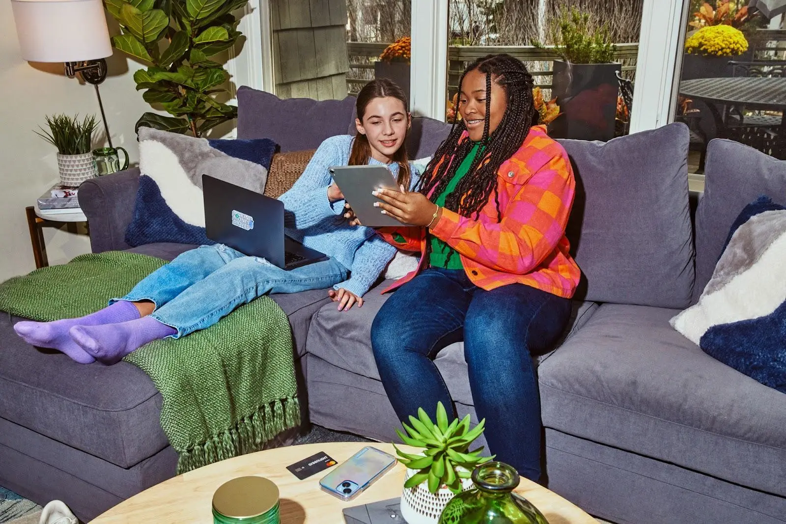 Two girls on a couch with a tablet and a computer chatting.