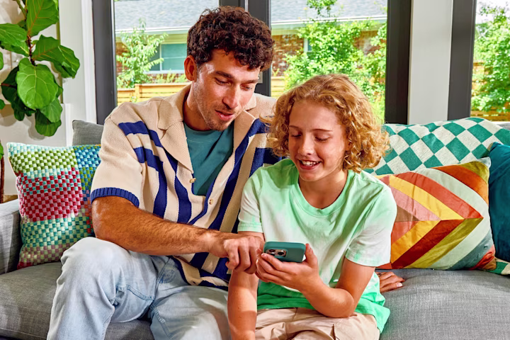 Adult and child looking at smartphone together on colorful couch with bright pillows and indoor plant nearby.