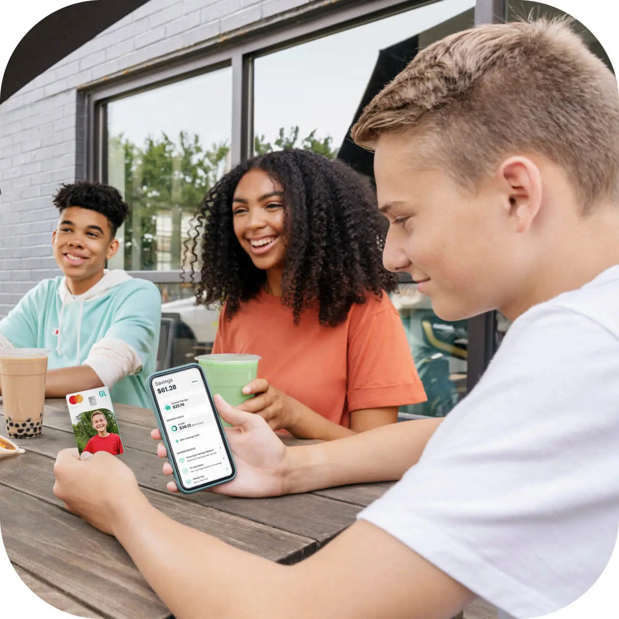 teenagers sitting at a table with one teen boy looking at his greenlight debit card and app