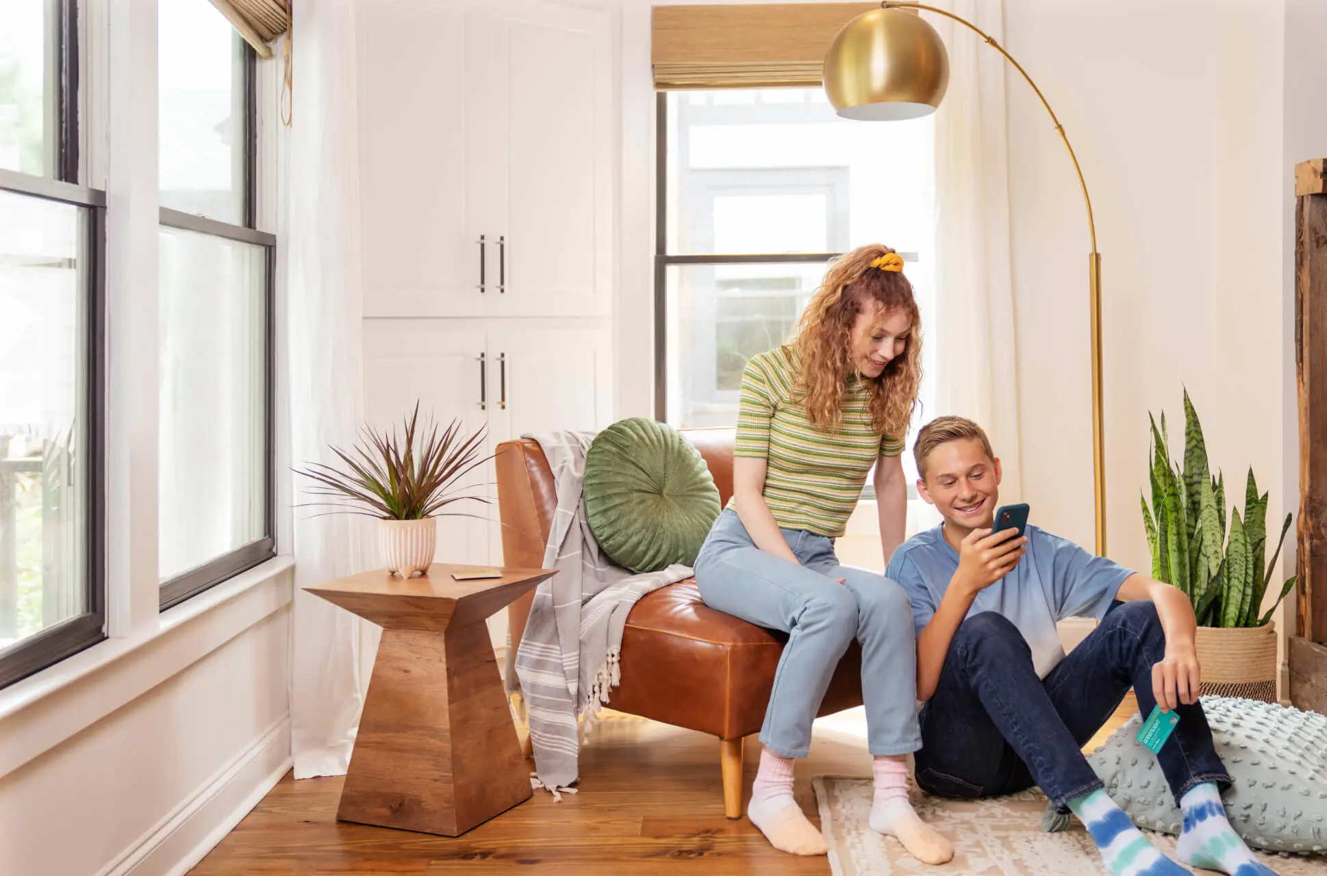 Teen girl and boy hanging out in their living room in the city looking at Greenlight mobile app with investing