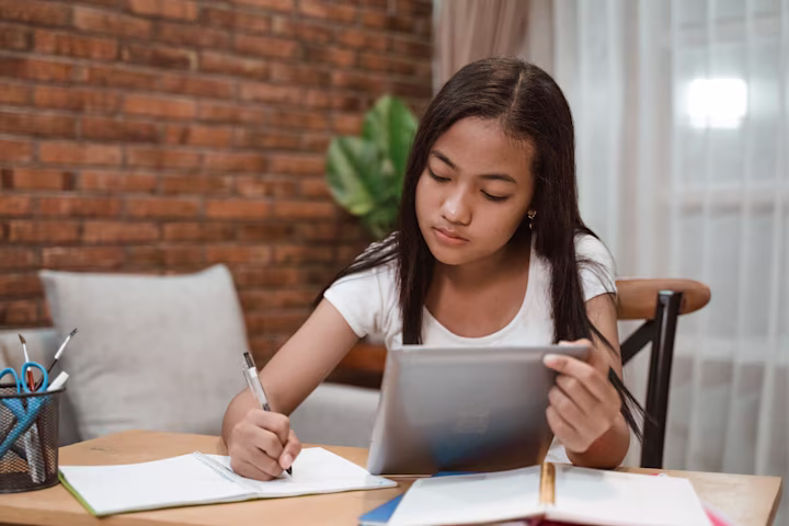 A young girl uses a tablet and writes in a notebook while sitting at a table