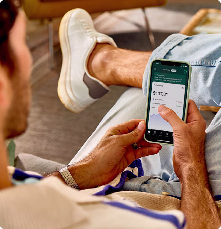Person in casual attire checking investment performance on smartphone showing $137.31 portfolio value with stock graphs.