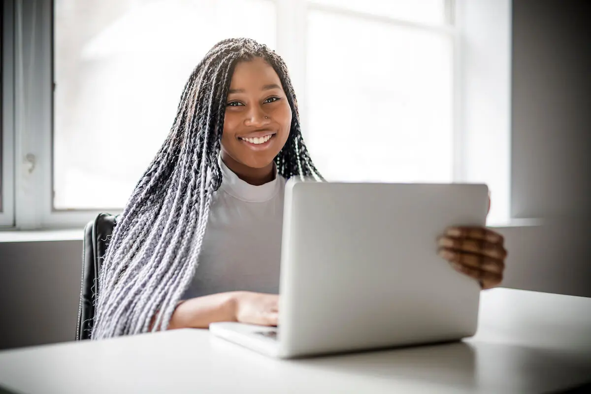 A smiling teenage girl sits at a table and uses a laptop