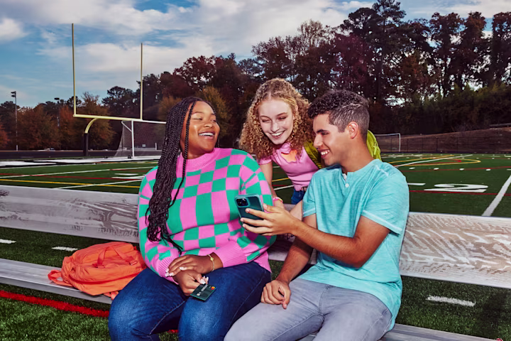Students sitting on football field bleachers looking at a phone together.