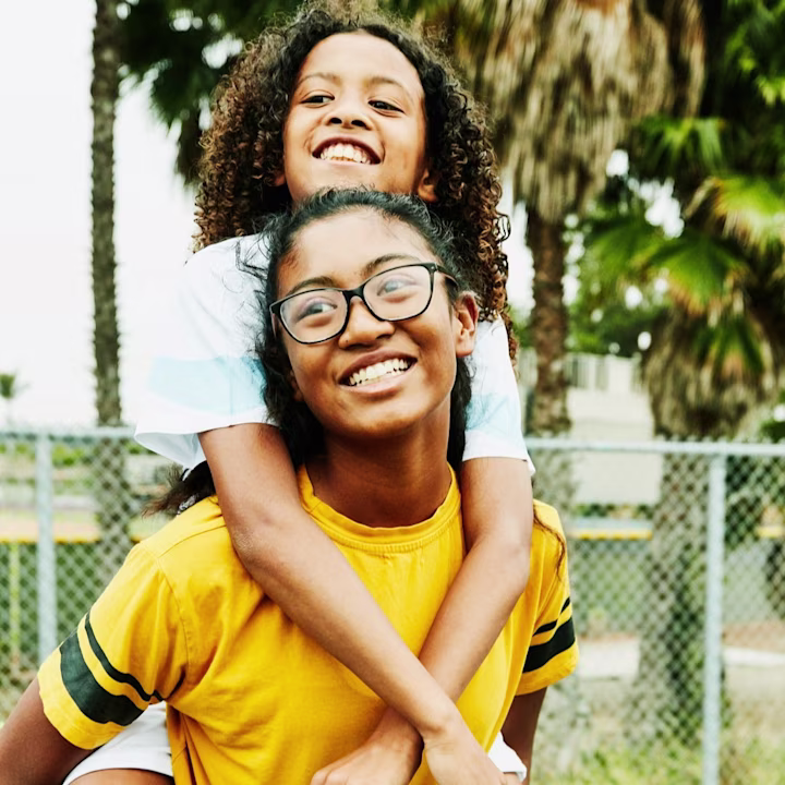 A girl riding piggy back on another girl outside on a field.