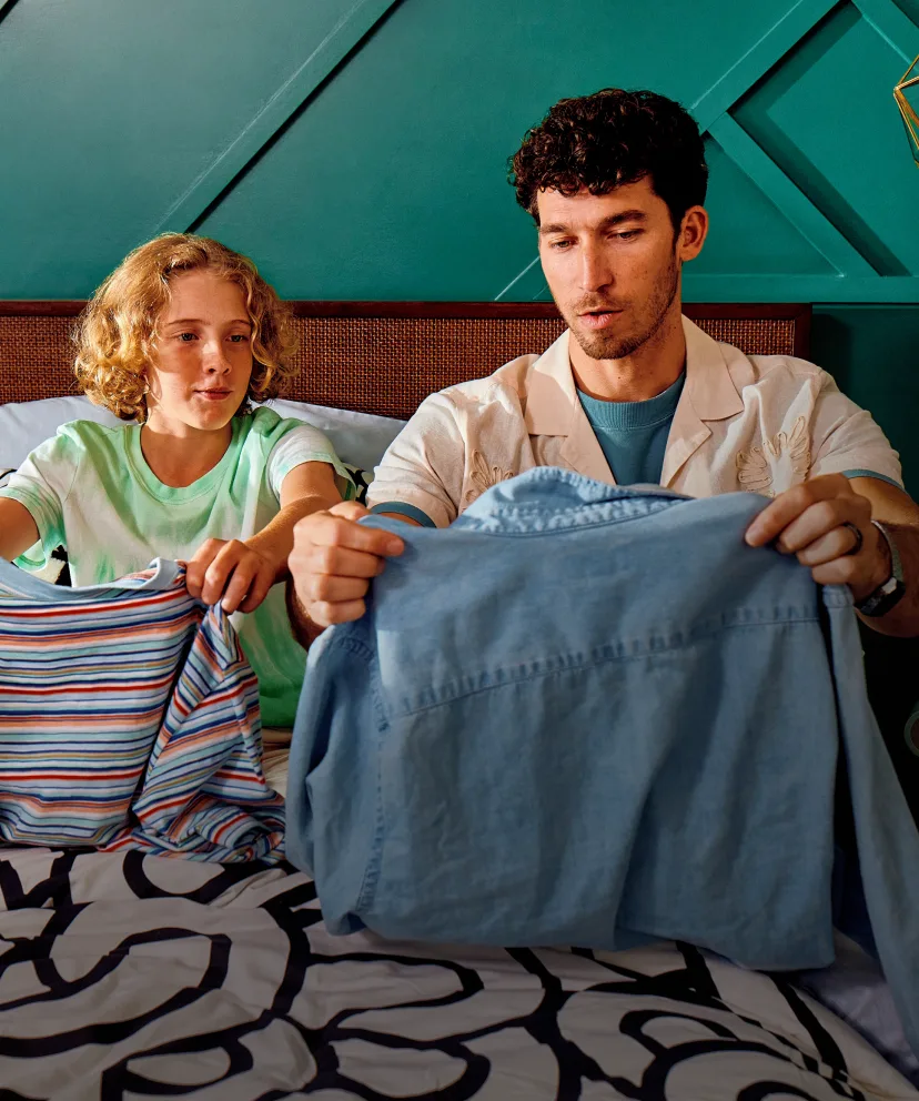 Two people sitting on a bed with teal wall background, folding blue and striped clothing items together.