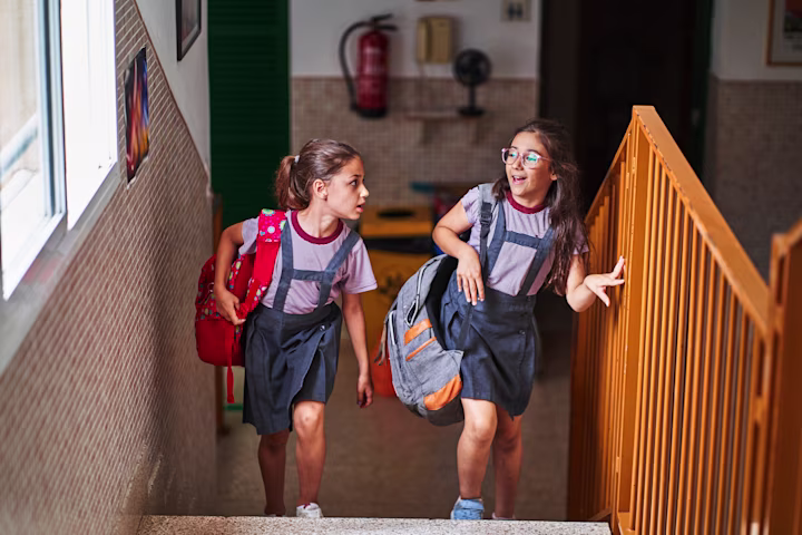 Two girls wearing backpacks and walking up stairs together at school.
