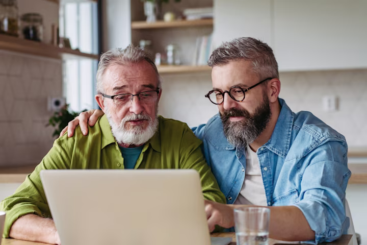 Two elderly men sitting at a kitchen table looking at a laptop together.