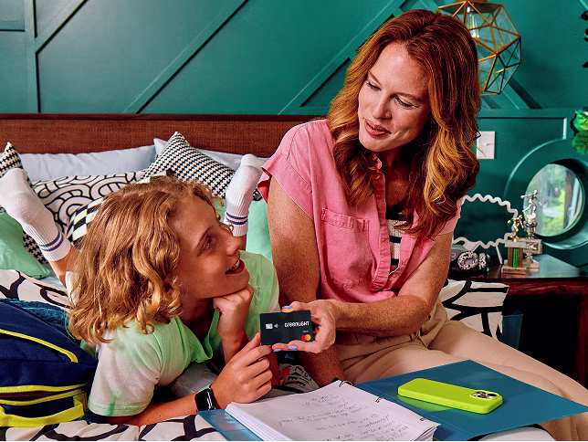 A mom shows her smiling child a Greenlight debit card as they relax on a bed.