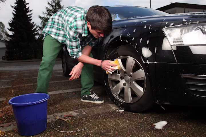 Teenage boy washes the wheels of a car with a bucket and sponge