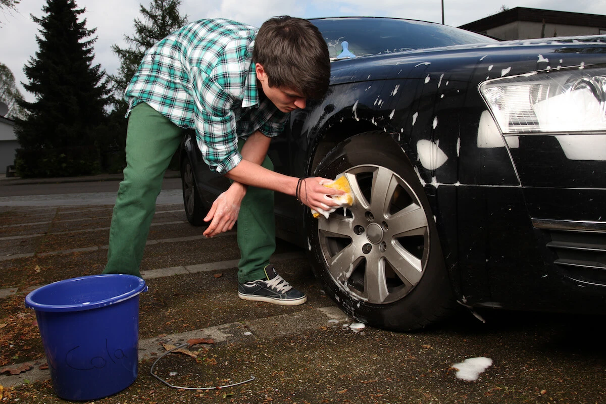 Small business ideas for teens: A teenage boy washes the wheels of a car with a bucket and sponge