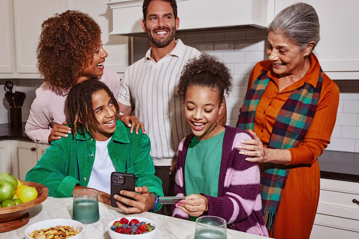 Parents, grandmother and teens in kitchen looking at phone.