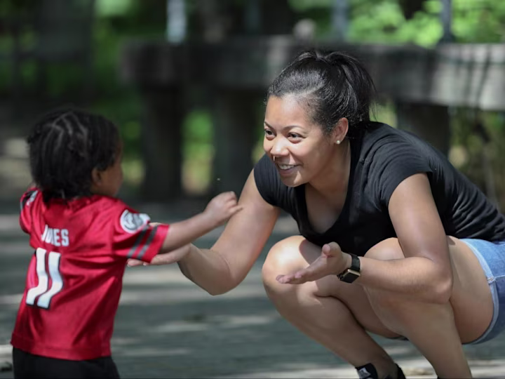 A mother kneeling down smiling outside as her child runs to her.