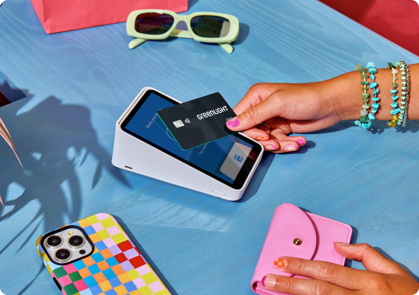Hand tapping Greenlight card on payment terminal with yellow sunglasses, colorful phone case, and pink wallet nearby.