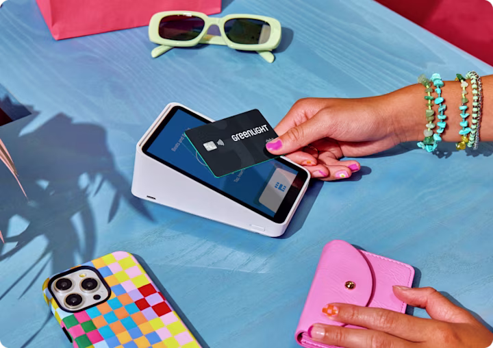 Hand tapping Greenlight card on payment terminal with yellow sunglasses, colorful phone case, and pink wallet nearby.