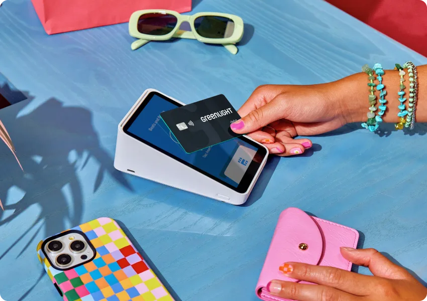 Hand tapping Greenlight card on payment terminal with yellow sunglasses, colorful phone case, and pink wallet nearby.