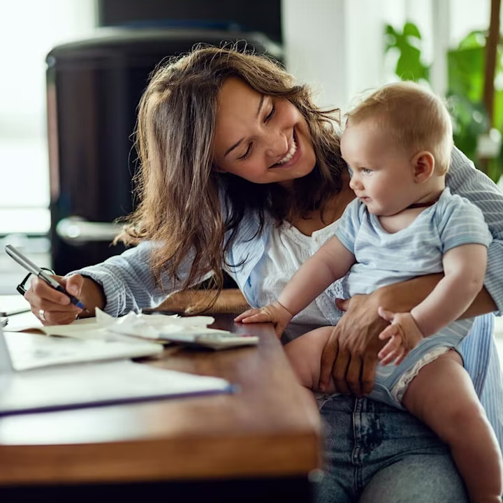 A smiling mom holds her baby on her lap while she looks through bills and receipts