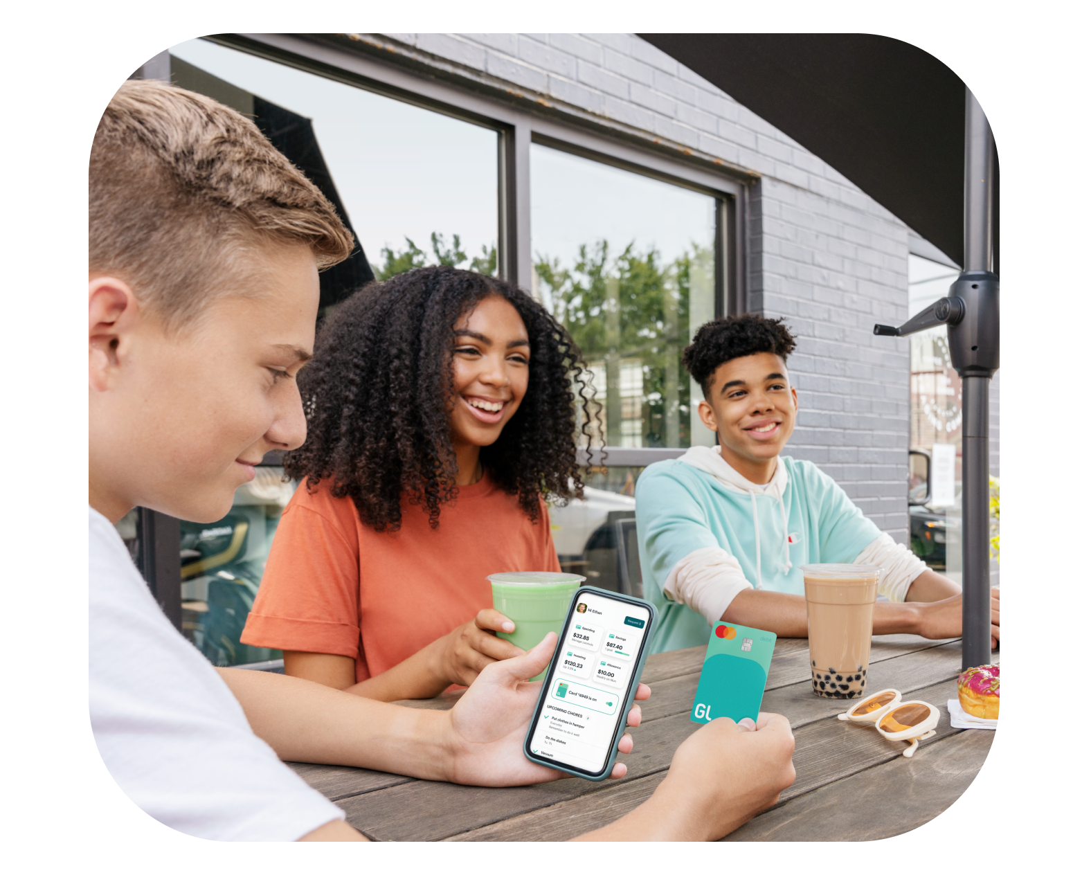 Three teens sitting outside laughing while one teen uses Greenlight debit card and mobile app to refer a friend and collect rewards