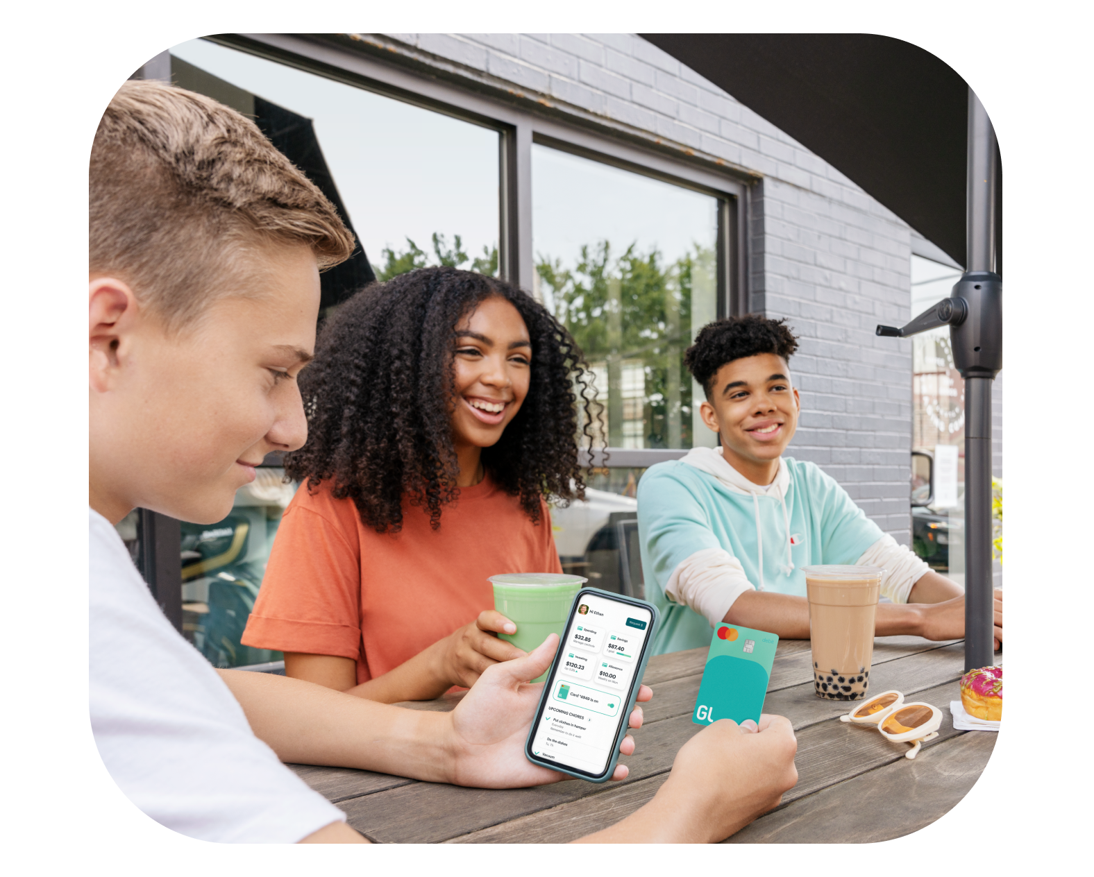 Three teens sitting outside laughing while one teen uses Greenlight debit card and mobile app to refer a friend and collect rewards
