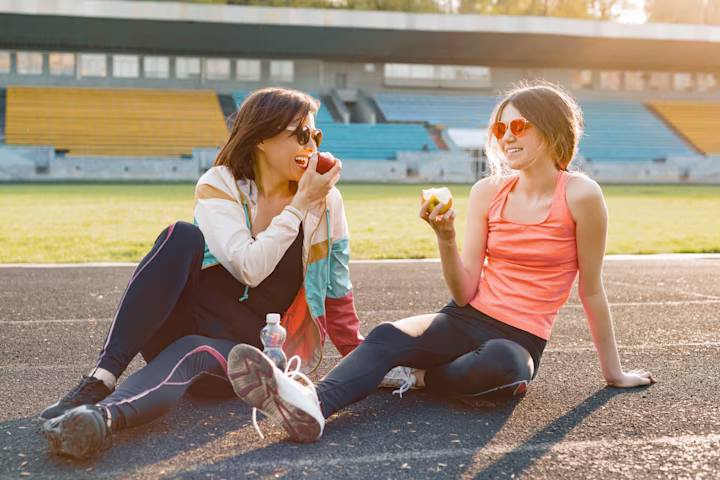 Two women sit on a track together eating a snack.