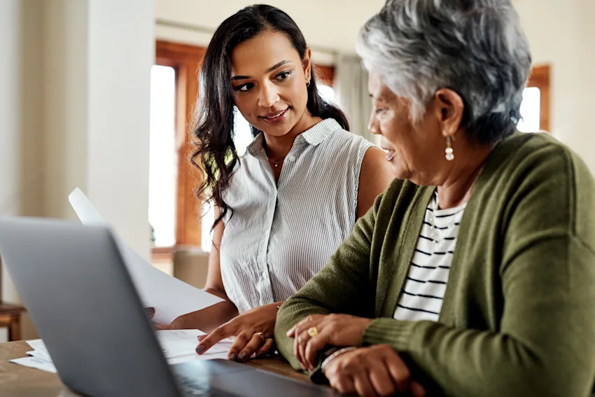 Daughter and elderly mother looking at paperwork while working on a laptop together.