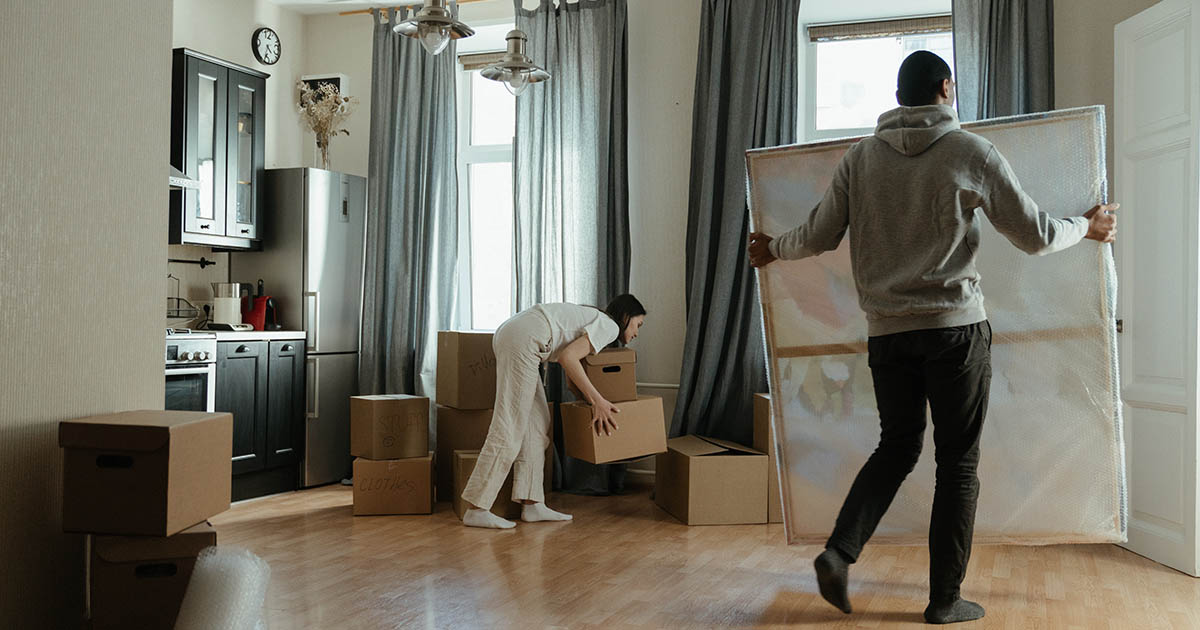 Ett par flyttar in i bostad, kvinnan packar upp kartonger i köket och mannnen bär en stor tavla.