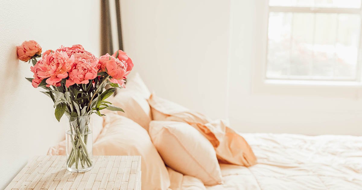 Säng med rosa lakan och vita väggar i bakgrunden, samt ett nattduksbord med en bukett rosa blommor på.