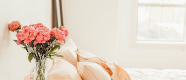 Säng med rosa lakan och vita väggar i bakgrunden, samt ett nattduksbord med en bukett rosa blommor på.